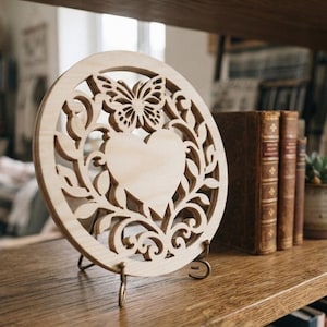 May include: A circular light-colored wooden cutout featuring an intricate laser-cut design of a central heart surrounded by swirling vine patterns and a butterfly at the top. The piece is displayed upright on a small metal stand.