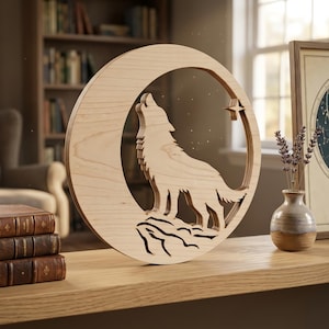 Pode incluir: Um recorte circular de madeira de cor clara apresentando a silhueta de um lobo uivando para uma lua crescente. O design inclui uma pequena estrela de quatro pontas e recortes intrincados representando terreno rochoso na base do lobo.