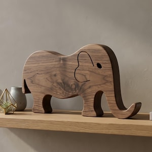 May include: A wooden elephant figurine with a curved trunk and large ears. The elephant is made of a dark brown wood and is displayed on a light-colored wooden shelf. The figurine has a simple, minimalist design and is a decorative item.