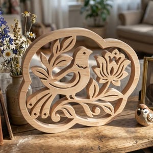 Puede incluir: Escultura decorativa de madera que representa un pájaro posado en una rama con hojas y una flor. La talla de madera de color claro está enmarcada en una forma redondeada. El diseño está inspirado en la naturaleza y está sobre una superficie de madera.