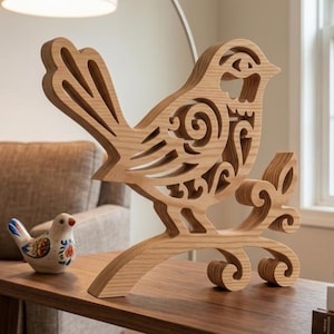 May include: A light-colored wooden bird sculpture featuring intricate cut-out scrollwork patterns, perched on a stylized branch base. Beside it sits a small, smooth ceramic bird figurine decorated with colorful floral patterns in blue, red, and green.