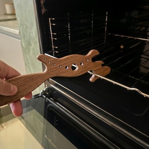 Puede incluir: Un tirador de rejilla de horno de madera con un diseño estilizado. El tirador es de madera marrón claro y presenta una serie de pequeños recortes circulares. Se utiliza para sacar una rejilla del horno.