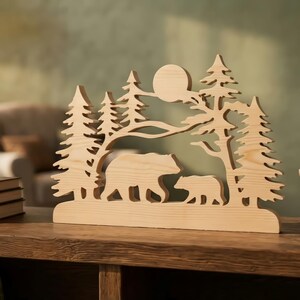Puede incluir: Un recorte de madera de color claro que presenta una escena forestal con un oso grande y un osezno caminando entre pinos bajo una luna circular. La pieza decorativa tiene una base plana y muestra la veta natural de la madera clara.