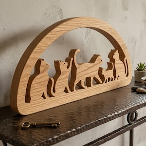 May include: A wooden cat silhouette sculpture featuring a row of stylized cat figures within an arched frame. The sculpture is made of light-coloured wood and sits on a dark metal surface. A vintage key rests on the surface.