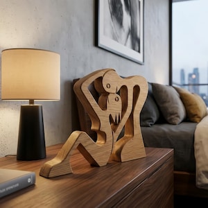 May include: A light brown wooden sculpture depicting two figures in an embrace. The sculpture is placed on a dark brown wooden surface. A black lamp with a beige lampshade is positioned to the left of the sculpture.