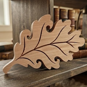 May include: A light brown wooden cutout shaped like an oak leaf with smooth edges and deep, carved vein details running through the center and extending toward the lobed edges of the leaf, showcasing a natural wood grain texture throughout.