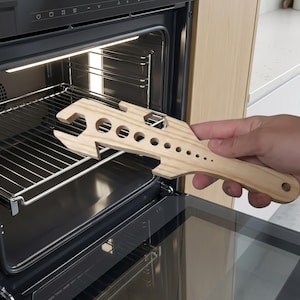 May include: A wooden oven rack puller with a wrench-like design. The tool has multiple circular cutouts and a handle. The puller is being used to pull out the oven rack. The tool is made of light-colored wood.