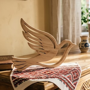 Könnte beinhalten: Eine Holzskulptur einer fliegenden Taube aus hellem Holz. Die Flügel des Vogels sind ausgebreitet, und das Design weist einen Ausschnittstil auf. Die Skulptur steht auf einem Tisch mit einem gemusterten Textil, was auf einen Dekorationsartikel hindeutet.