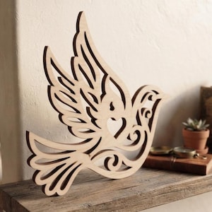 May include: A light-colored wooden cutout of a bird in flight with intricate scrollwork patterns and a heart-shaped cutout in the center of its body. The bird is depicted with wings spread upward and a fanned tail, featuring smooth, clean-cut edges.