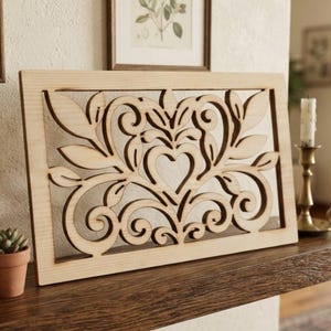 May include: A rectangular light-colored wooden panel featuring an intricate laser-cut design. The pattern consists of symmetrical scrolling flourishes, stylized leaves, and a central heart shape, all framed within a solid wood border.