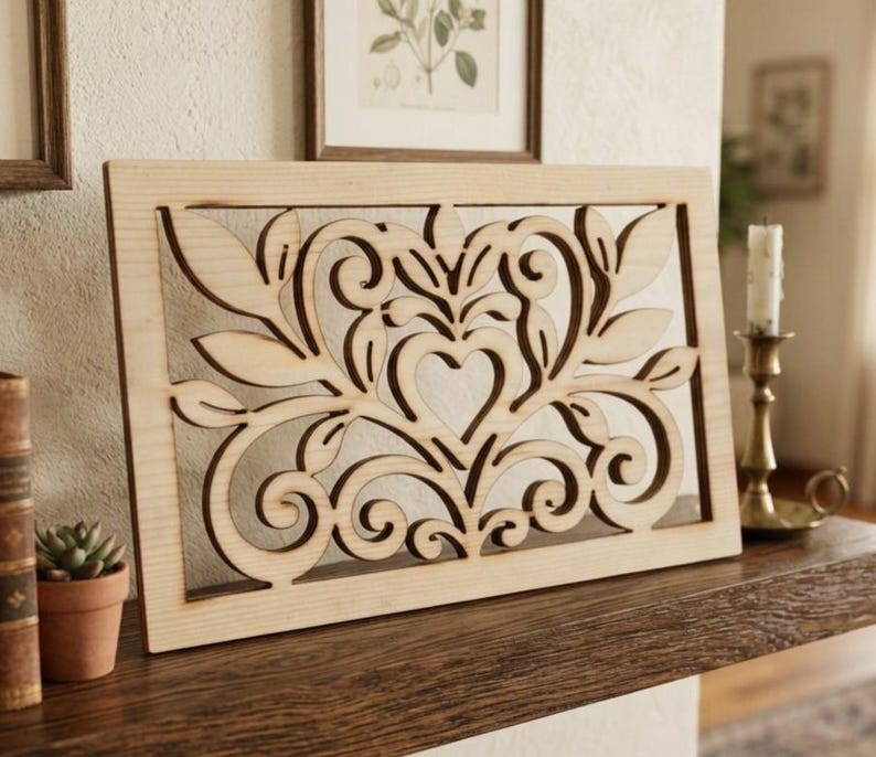May include: A rectangular light-colored wooden panel featuring an intricate laser-cut design. The pattern consists of symmetrical scrolling flourishes, stylized leaves, and a central heart shape, all framed within a solid wood border.