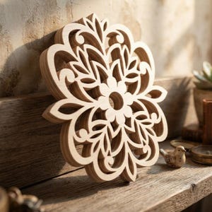 Può includere: Un elemento decorativo da parete in legno chiaro caratterizzato da un motivo floreale a mandala simmetrico e complesso tagliato al laser, con forme curve a petalo e un'apertura circolare centrale, che mette in risalto la venatura naturale.
