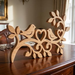 Könnte beinhalten: Dekorative Holzskulptur mit zwei Vögeln auf einem Ast mit herzförmigen Ausschnitten. Die Skulptur ist aus hellem Holz gefertigt und hat eine natürliche Oberfläche. Die Vögel und Äste sind aufwendig geschnitzt und schaffen ein charmantes und elegantes Stück.