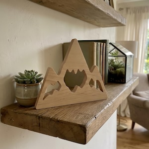 Puede incluir: Un adorno de estante en forma de montaña de madera clara con detalles recortados, colocado sobre una estantería de madera rústica. Una pequeña suculenta en una maceta de cerámica y un terrario de vidrio también están en el estante.