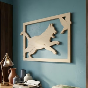May include: Wooden wall art featuring a cat chasing a bird within a rectangular frame. The cat is in mid-leap, with its mouth open, and the bird is positioned in the upper right corner. The artwork is made of light-colored wood and mounted on a blue wall.