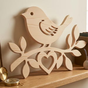 May include: A light-colored wooden wall decoration featuring a bird perched on a branch with leaves. The design includes a cutout heart shape integrated into the branch structure, showcasing the natural wood grain and smooth, flat-cut edges.
