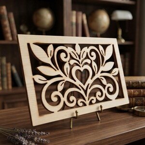 May include: A rectangular light-colored wooden panel featuring an intricate laser-cut design of symmetrical swirling vines, leaves, and a central heart shape. The panel is supported by a small metal stand, displaying a detailed cutout pattern.