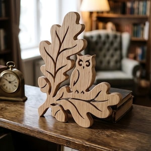 May include: A light brown wooden decorative piece featuring a carved owl perched on a large oak leaf. The natural wood grain is visible throughout the cutout design, which displays a detailed silhouette of the bird and the leaf structure.