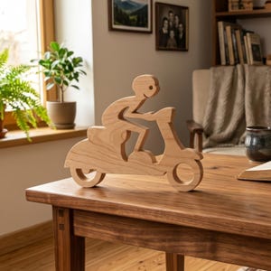 Puede incluir: Escultura de madera de una persona montada en un scooter. La escultura es de madera clara y presenta un diseño minimalista. El scooter tiene una rueda delantera redonda y un asiento. La persona tiene una cara simple y sonriente.