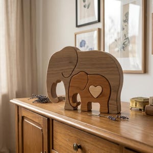 May include: A wooden elephant family sculpture. The large elephant is light brown, with a smaller, medium-brown elephant nested inside. The small elephant has a heart-shaped cutout. The sculpture is displayed on a wooden dresser.