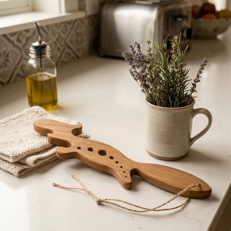 May include: A light brown wooden herb stripper tool with a long, curved handle and a series of circular holes of varying sizes along its center. A thin, light brown twine string is attached to the end of the wooden tool for hanging.