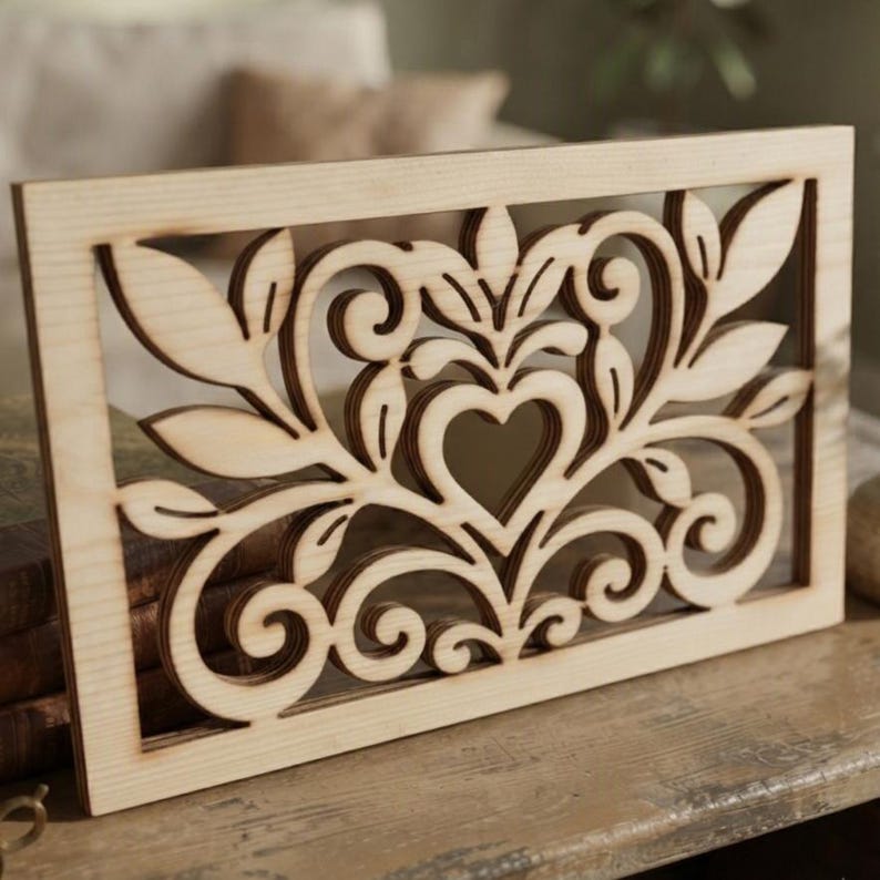 May include: A rectangular light-colored wooden panel featuring an intricate laser-cut design. The pattern consists of symmetrical scrolling vines, stylized leaves, and a central heart-shaped cutout, all framed within a solid wood border.