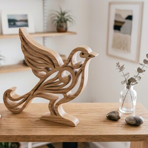 May include: A light brown wooden bird sculpture featuring intricate cut-out patterns throughout its body and wings. The bird is depicted in a standing position with its wings partially extended, showcasing the natural wood grain and smooth finish.