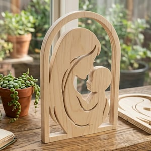 Può includere: Scultura in legno raffigurante una madre e un bambino all'interno di una cornice ad arco. La venatura naturale del legno è visibile. La madre culla il bambino, entrambe le figure sono di profilo. La scultura è su un tavolo di legno vicino a una finestra con piante.