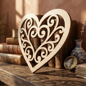 May include: A light-colored wooden heart cutout featuring an intricate, symmetrical scrollwork pattern throughout its interior. The heart is positioned upright, displaying a smooth, flat surface and detailed cutouts that form a decorative design.