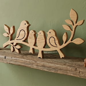 Puede incluir: Decoración de pared de madera que representa tres pájaros posados en una rama con hojas. Los pájaros y la rama están hechos de madera clara, con detalles grabados. La obra de arte está montada en una estantería de madera rústica contra una pared verde.