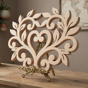 May include: A light-colored wooden cutout featuring an intricate, symmetrical scrollwork design with leaf-like shapes and a central heart-shaped opening. The wooden piece is supported by an ornate, metallic gold-toned display stand with curved legs.