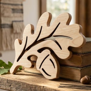 May include: A wooden oak leaf and acorn decoration. The light-colored wood is carved with detailed veins and outlines. The leaf is resting on a stack of old books, with a natural, rustic aesthetic.
