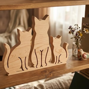 May include: Wooden cat silhouette decor featuring three cats of varying sizes. The light-colored wood has visible grain and is placed on a wooden shelf. A small glass jar with flowers and a ceramic bird are also on the shelf.