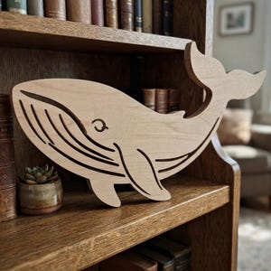 May include: A light-coloured wooden cutout of a whale featuring engraved lines to represent the throat pleats, a simple eye, and a curved fin. The whale figure is oriented horizontally with its tail raised, showcasing a smooth, natural wood grain finish.