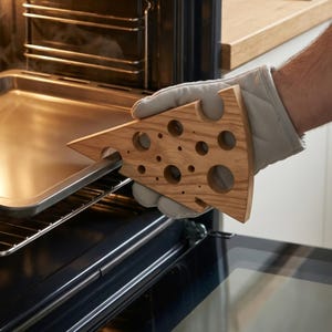 Peut inclure: Un tire-plat en bois en forme de morceau de fromage, avec diverses découpes circulaires. Le tire-plat est utilisé pour retirer une plaque de cuisson métallique d'un four. La personne porte un gant de cuisine gris.