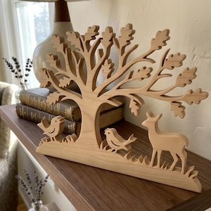 May include: A light-colored wooden cutout featuring a central tree with detailed oak leaves, two small birds perched on the base, and a standing deer. The smooth, natural wood sculpture is crafted as a single piece with a flat, stable base.