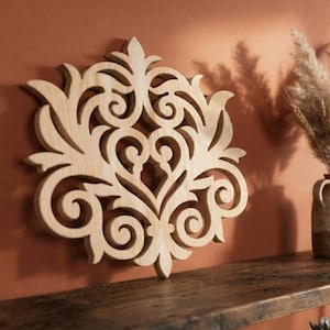 May include: A light-colored wooden wall decoration featuring an intricate, symmetrical laser-cut design with swirling scrollwork and leaf-like patterns. The piece has a flat, layered appearance with a natural wood grain texture and smooth edges.