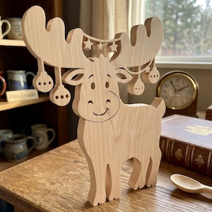 May include: A light-colored wooden moose figurine featuring a cutout design of antlers decorated with hanging circular ornaments and small stars. The moose has a smooth, natural wood grain finish, a smiling face, and stands upright on a flat base.