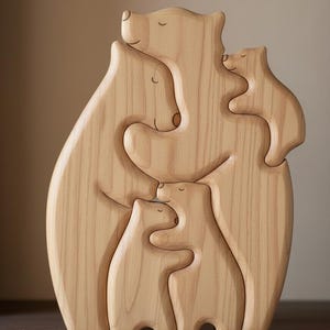 Puede incluir: Una escultura de madera de una familia de osos. La escultura presenta un oso grande abrazando a dos osos más pequeños, con dos osos adicionales anidados en su interior. Los osos están tallados en madera clara, con bordes lisos y redondeados.