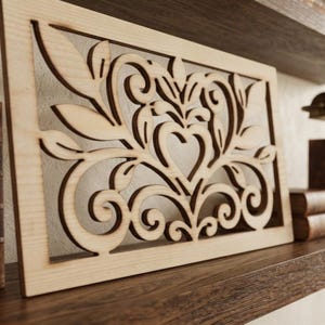 May include: A rectangular light-colored wooden panel featuring a laser-cut design of symmetrical swirling vines, leaves, and a central heart shape. The intricate cutout pattern is framed by a solid wood border with a smooth, natural grain finish.