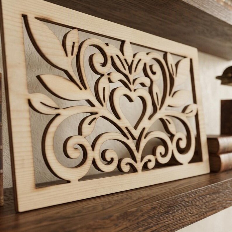 May include: A rectangular light-colored wooden panel featuring a laser-cut design of symmetrical swirling vines, leaves, and a central heart shape. The intricate cutout pattern is framed by a solid wood border with a smooth, natural grain finish.