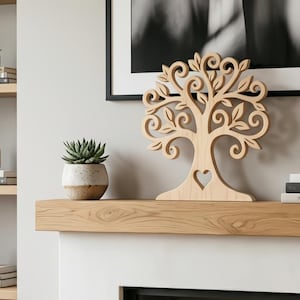 May include: A light-colored wooden tree sculpture with a heart-shaped cutout in the trunk. The tree has intricate leaf and swirl details. It is displayed on a wooden mantel with a potted succulent and a framed black and white photograph.