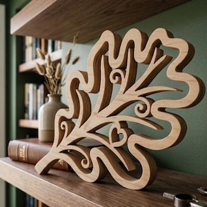May include: A light brown wooden cutout in the shape of an oak leaf with intricate scrollwork and a small heart detail integrated into the design. The object features a smooth, natural wood grain finish and a layered, open-work structure.