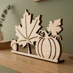 May include: A light-colored wooden cutout decoration featuring a large maple leaf, a pumpkin, and a small acorn. The flat, laser-cut design is mounted on a rectangular base, showcasing the natural wood grain and dark edges from the cutting process.