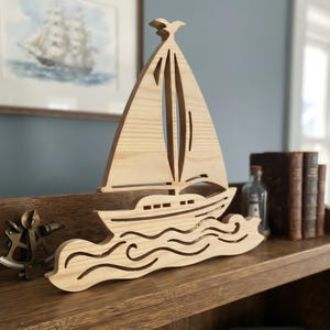 May include: A light-colored wooden cutout of a sailboat featuring a tall mast, a triangular sail, and a hull resting on stylized, wavy water. The design is crafted from natural wood with visible grain patterns, showcasing a clean, flat silhouette.