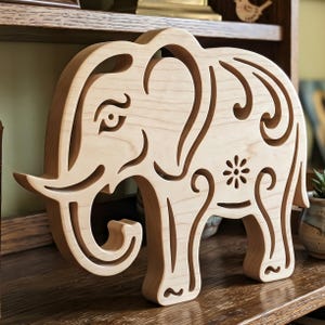 May include: A light-colored wooden elephant sculpture with intricate cut-out details. The elephant features a stylized eye, decorative swirls, and a floral design. The sculpture is displayed on a wooden shelf.