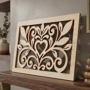 May include: A rectangular light-colored wooden panel featuring an intricate laser-cut design. The pattern consists of symmetrical swirling scrolls, stylized leaves, and a central heart-shaped cutout, all framed within a solid wood border.