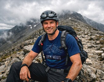 May include: A man wearing a helmet and sunglasses, a blue t-shirt with a mountain graphic and the text "BEN NEVIS RIDGE SPECIALIST", and a backpack, sits on a rocky mountain ridge. The background features a cloudy sky and mountain range.