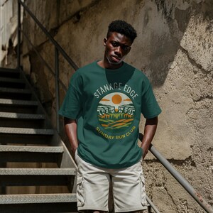 May include: A dark green t-shirt with a circular graphic that reads "STANAGE EDGE SUNDAY RUN CLUB". The design includes a sun, hills, and trees. The person is wearing khaki cargo shorts.