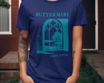 May include: A navy blue t-shirt featuring a teal graphic of a gothic window overlooking a landscape. The text "BUTTERMERE" is above the window, and "DAWN DIPPERS" is below.